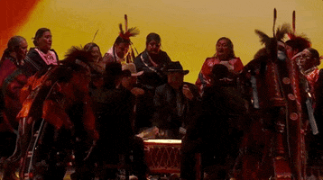 Oscars 2024 gif. Scott George and Osage singers perform 'Wahzhazhe (A Song For My People)' from Killers of the Flower Moon. While an inner circle of men play a powwow drum, a group of singers in Indigenous wear sing and dance around them. 