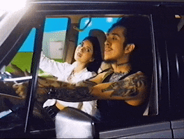 Music video gif. In a scene from the music video "Cuco" Aleman sits in the driver's seat of a car with a woman in the passenger seat. He sways his head as he sings and she looks over at him longingly. They are passing by a fake colorful background showing door after door of a perfect suburban neighborhood. 