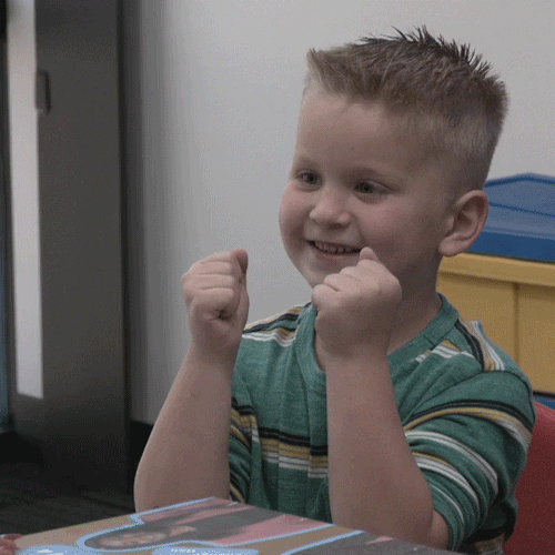 Video gif. A beaming young boy sits at a table with his hands clenched. His smile grows into an excited face as he feverishly interlocks his fingers.
