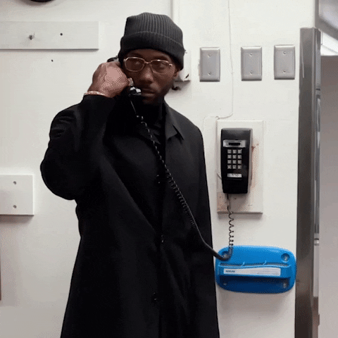 Video gif. NBA player Kawhi Leonard in a black suit, shirt, and beanie picks up a phone on the wall of a backstage area, looking out at someone with a suspicious stare. 