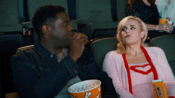 Movie gif. Rebel Wilson as Stephanie and Sam Richardson as Seth in Senior Year. They're in a movie theater and Seth tries to shoot popcorn into Stephanie's mouth. He fails and they both chuckle.
